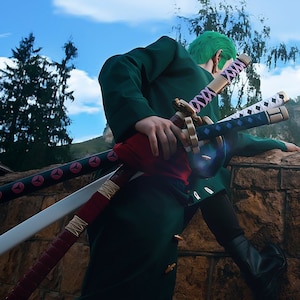 May include: A person dressed as a character from the anime One Piece, with green hair and a green jacket, holding three swords. The swords have different colored wrappings and designs. The person is standing on a stone wall, looking out at a landscape.