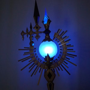 May include: A decorative metal staff with a glowing blue orb at the center, surrounded by a radiating sunburst design. The staff has a pointed top and a base with intricate details.