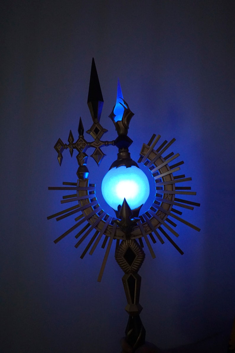 May include: A decorative metal staff with a glowing blue orb at the center, surrounded by a radiating sunburst design. The staff has a pointed top and a base with intricate details.