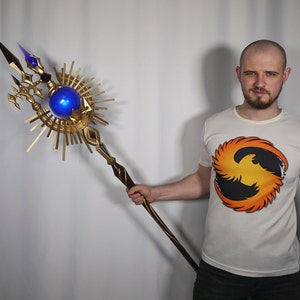May include: A person holding a gold and blue staff with a large orb at the top. The staff has a wooden handle and a decorative design. The person is wearing a white t-shirt with a phoenix design.