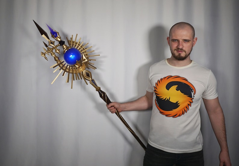 May include: A person holding a gold and blue staff with a large orb at the top. The staff has a wooden handle and a decorative design. The person is wearing a white t-shirt with a phoenix design.