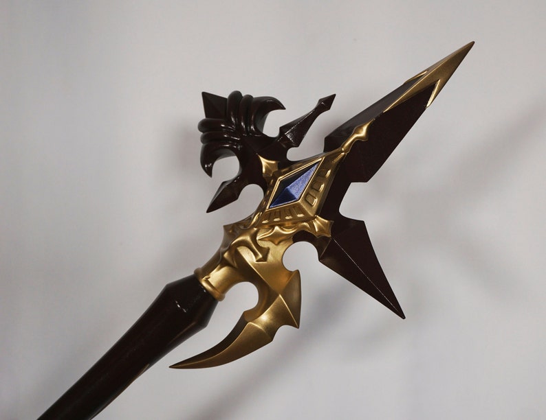 May include: A decorative spear with a gold and brown finish. The spear head features a detailed design with a blue gem in the center.