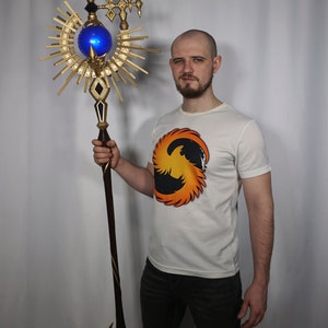 May include: A person holding a gold and blue staff with a large, ornate, sun-shaped design. The staff has a blue orb at the top and a sharp point at the bottom.