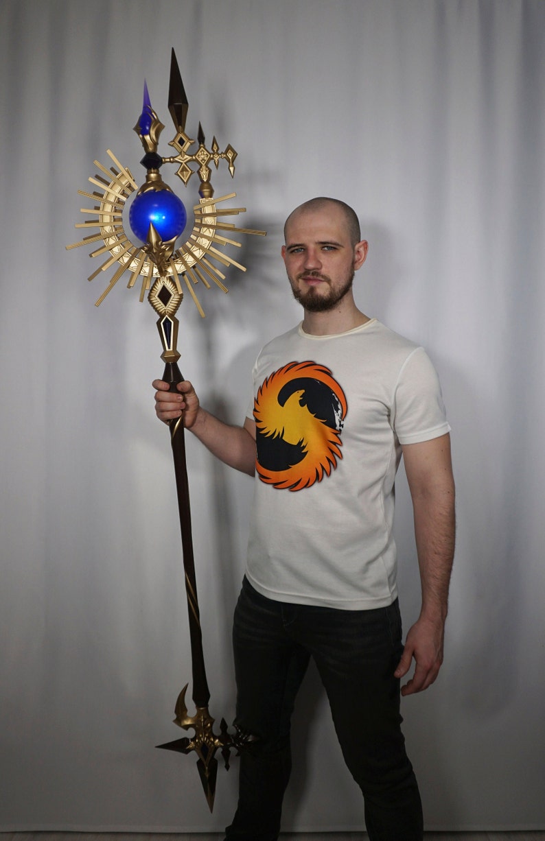May include: A person holding a gold and blue staff with a large, ornate, sun-shaped design. The staff has a blue orb at the top and a sharp point at the bottom.