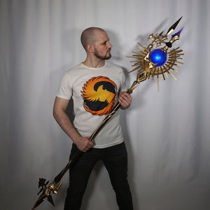May include: A person holding a large, ornate staff with a golden base and a blue orb at the top. The staff has a sunburst design and a sharp point at the end.