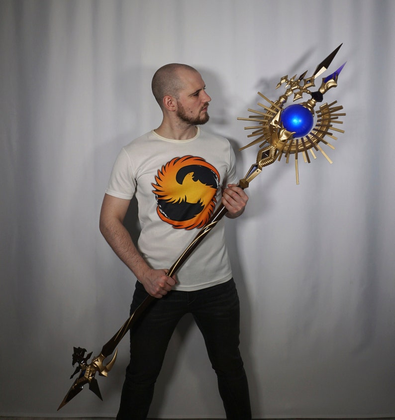 May include: A person holding a large, ornate staff with a golden base and a blue orb at the top. The staff has a sunburst design and a sharp point at the end.