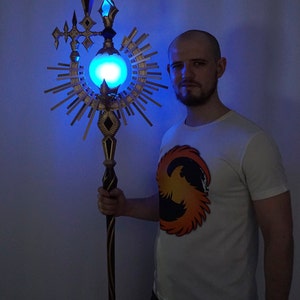May include: A person holding a staff with a glowing blue orb at the top. The staff is decorated with gold and black accents and has a sunburst design around the orb.