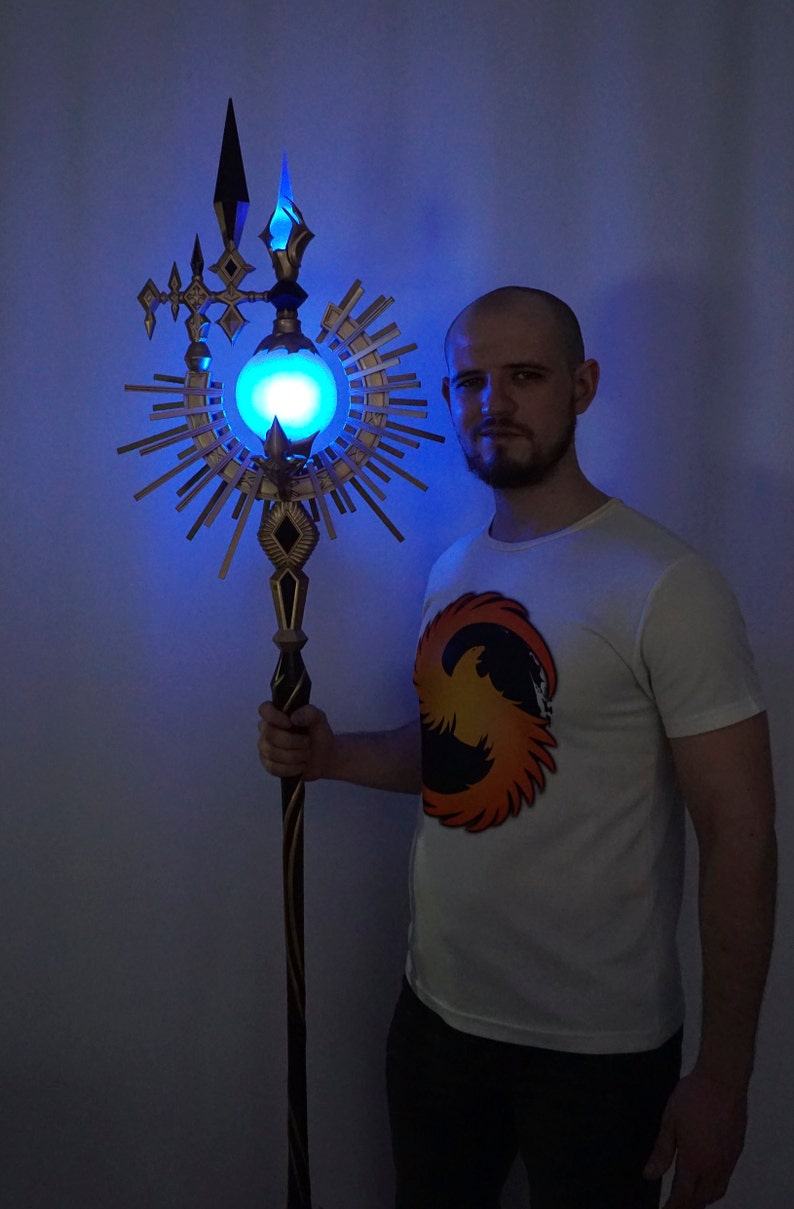 May include: A person holding a staff with a glowing blue orb at the top. The staff is decorated with gold and black accents and has a sunburst design around the orb.