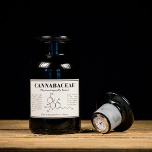 Pode incluir: Uma garrafa de vidro azul escuro com um rótulo preto que diz "CANNABACEAE Physiocologically Tested" e um diagrama químico. A garrafa tem uma tampa prateada e preta com um termômetro no lado. A garrafa está sobre uma superfície de madeira.