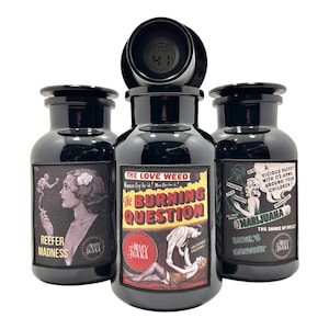 Può includere: Tre barattoli di vetro nero con etichette in stile vintage. Le etichette presentano testo e illustrazioni relative alla marijuana, tra cui "Reefer Madness", "The Burning Question" e "Marijuana". Un termometro digitale è posto sopra il barattolo centrale.