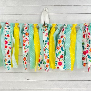 May include: A colorful fabric garland with yellow, blue, and white floral patterns. The garland is made of strips of fabric tied together and hangs from a white hook.