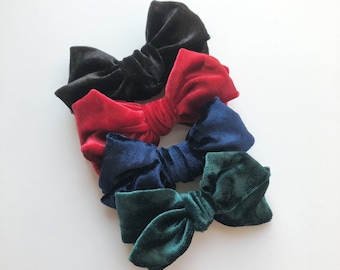 May include: Four velvet hair bows in black, red, navy blue, and forest green. The bows are arranged diagonally, showcasing their soft texture and rich colours. These accessories are ideal for adding a touch of elegance to any hairstyle.