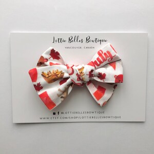 May include: A red, white and brown bow with a Canadian flag and maple leaf design. The bow is on a white card with the text "Lottie Belles Boutique, Vancouver, Canada" and social media handles.