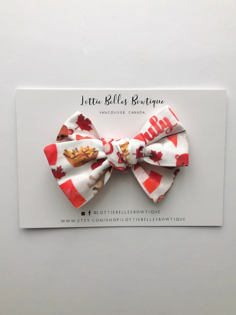 May include: A red, white and brown bow with a Canadian flag and maple leaf design. The bow is on a white card with the text "Lottie Belles Boutique, Vancouver, Canada" and social media handles.