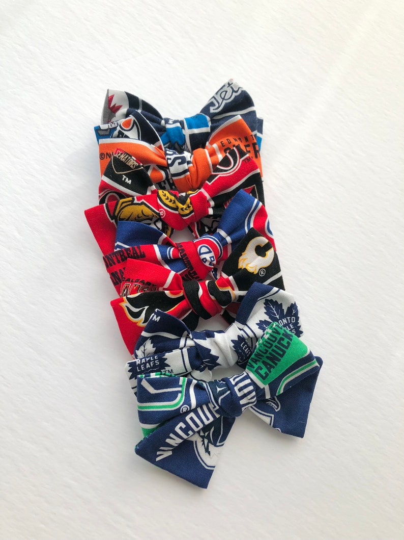 May include: Five different hockey team hair bows. The bows are made of fabric with the logos of the NHL teams: Edmonton Oilers, Montreal Canadiens, Calgary Flames, Toronto Maple Leafs, and Vancouver Canucks.