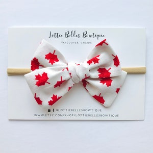May include: White headband with a red maple leaf patterned bow. The bow is tied in a knot and has a white background with red maple leaves. The headband is made of elastic and is suitable for babies and toddlers.