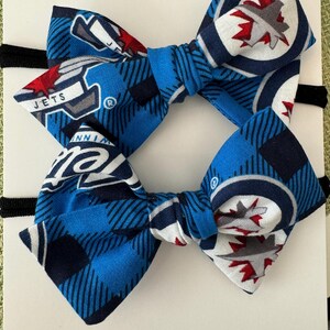 May include: Two blue and white bow headbands with a Winnipeg Jets hockey team logo. The bow is made of fabric with a blue and white plaid pattern. The logo is a white circle with a red maple leaf inside. The headbands are tied with black elastic.