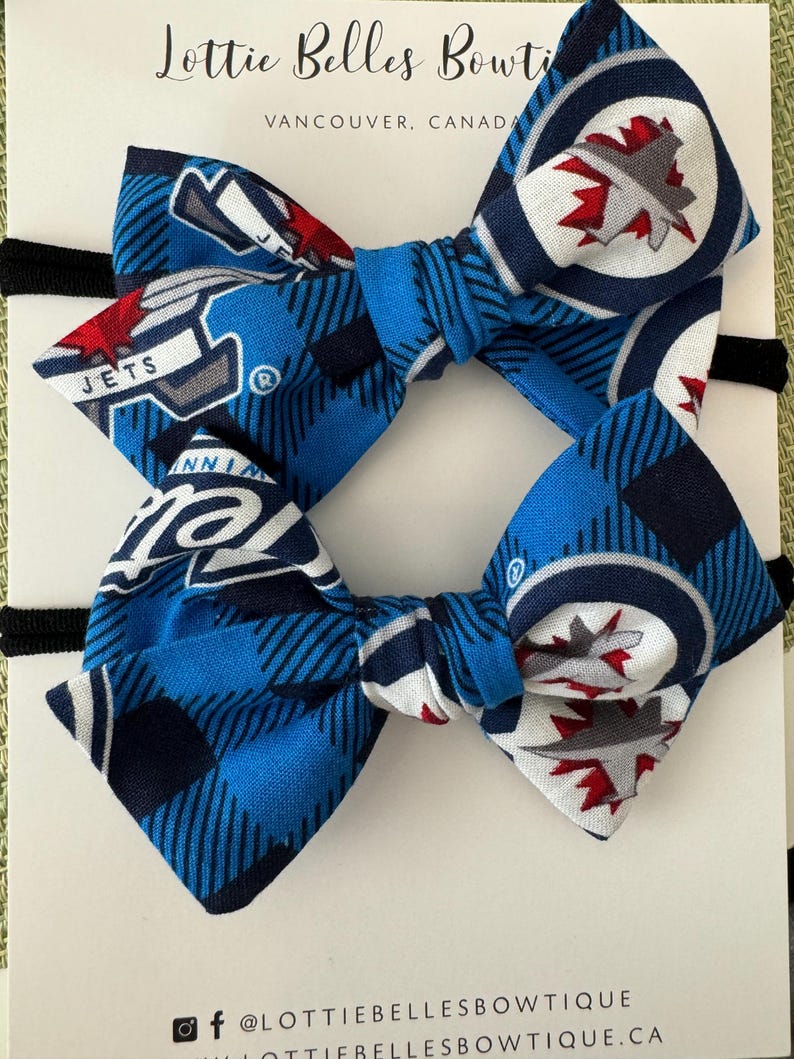 May include: Two blue and white bow headbands with a Winnipeg Jets hockey team logo. The bow is made of fabric with a blue and white plaid pattern. The logo is a white circle with a red maple leaf inside. The headbands are tied with black elastic.