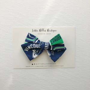 May include: A blue and green bow tie with the Vancouver Canucks logo. The bow tie is on a white card with the text "Lottie Belles Boutique, Vancouver, Canada" and social media information.
