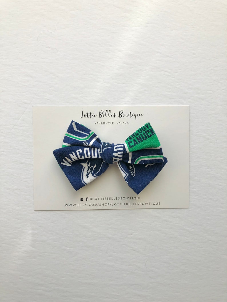 May include: A blue and green bow tie with the Vancouver Canucks logo. The bow tie is on a white card with the text "Lottie Belles Boutique, Vancouver, Canada" and social media information.