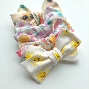 May include: Four fabric hair bows with different patterns. The bows are tied together and feature a white bow with yellow smiling suns on it.