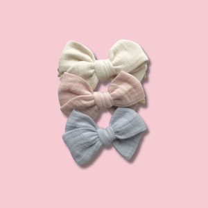 May include: Three fabric hair bows in cream, blush pink, and light blue, arranged vertically against a pink background. The bows are tied in a classic bow shape, with a textured, slightly wrinkled appearance. These accessories are suitable for babies and toddlers.