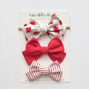 May include: Set of three hair bows with a Canadian theme. The first bow is white with red maple leaf print, the second is solid red, and the third is white with red stripes. The bows are displayed on a white card with the text "Lottie Belles Boutique Vancouver, Canada" and "www.lottie bellesboutique.com".
