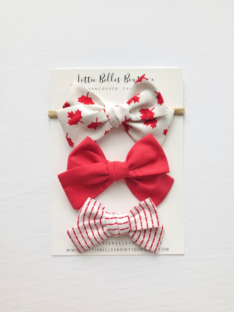 May include: Set of three hair bows with a Canadian theme. The first bow is white with red maple leaf print, the second is solid red, and the third is white with red stripes. The bows are displayed on a white card with the text "Lottie Belles Boutique Vancouver, Canada" and "www.lottie bellesboutique.com".