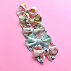 K&ouml;nnte beinhalten: F&uuml;nf Stoffhaarb&auml;nder mit rosa Hintergrund. Die Schleifen sind mit verschiedenen Disney-Prinzessinnenfiguren verziert.