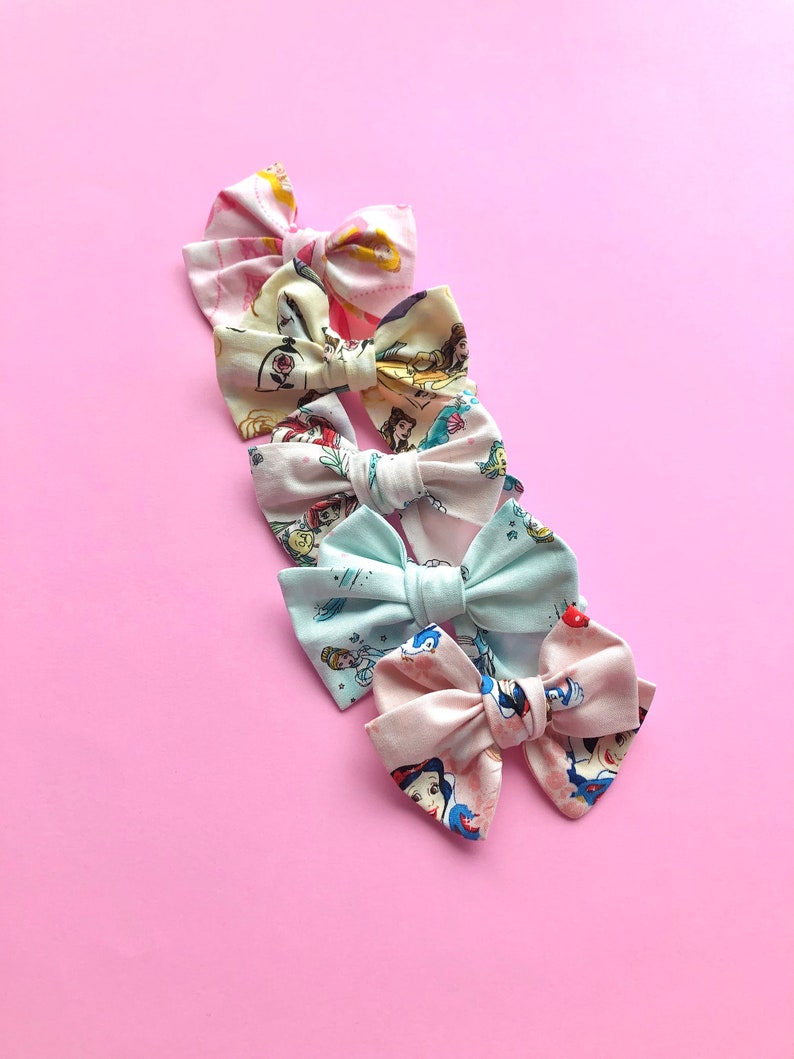 K&ouml;nnte beinhalten: F&uuml;nf Stoffhaarb&auml;nder mit rosa Hintergrund. Die Schleifen sind mit verschiedenen Disney-Prinzessinnenfiguren verziert.