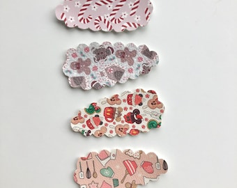 May include: Four decorative hair clips with scalloped edges. The clips feature various festive patterns, including candy canes, gingerbread men, and baking utensils, in shades of pink, red, and brown. The clips are arranged on a white background.