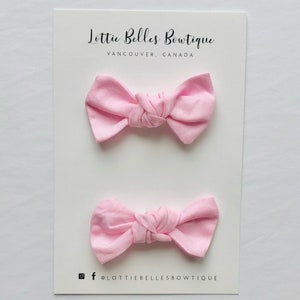 May include: Two pink fabric hair bows tied in a knot. The bows are on a white card with the text "Lottie Belles Boutique, Vancouver, Canada" and "Knot Pigtails".