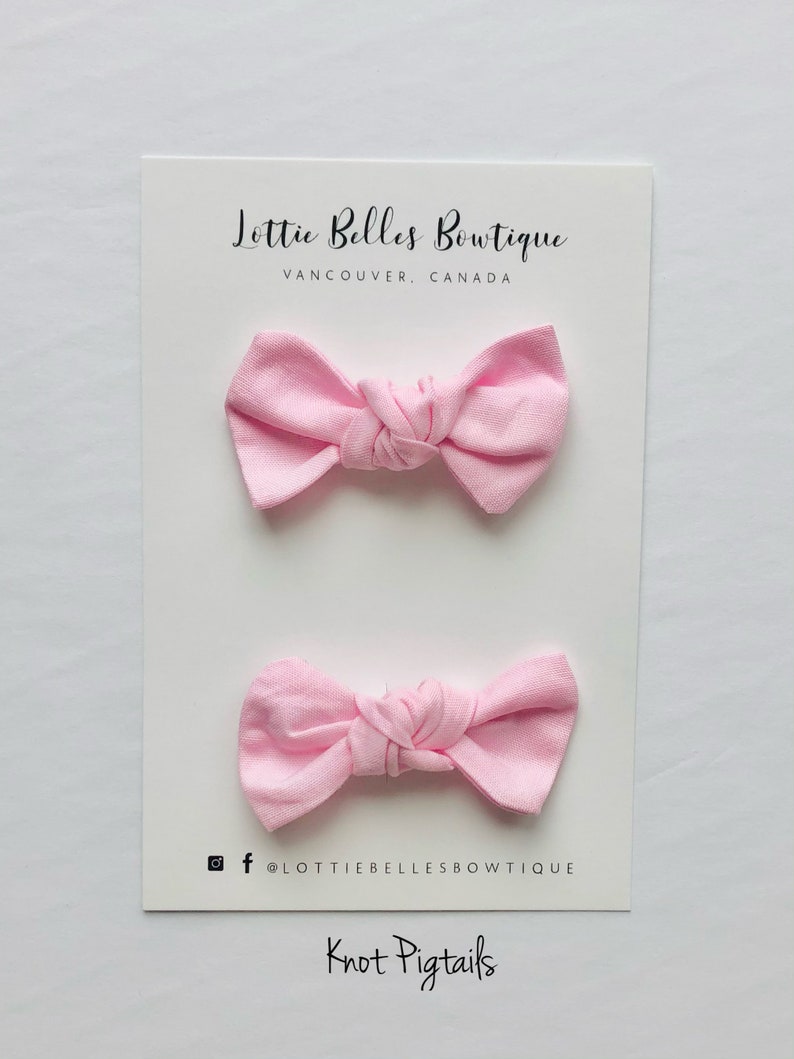 May include: Two pink fabric hair bows tied in a knot. The bows are on a white card with the text "Lottie Belles Boutique, Vancouver, Canada" and "Knot Pigtails".