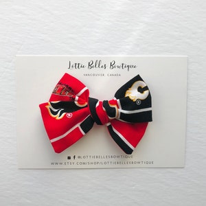 May include: A red, black, and yellow bow with a Calgary Flames and a  Edmonton Oilers hockey team logo. The bow is on a white card with the text "Lottie Belles Boutique, Vancouver, Canada" and the text "@lottie bellesboutique, www.etsy.com/shop/lottie bellesboutique".