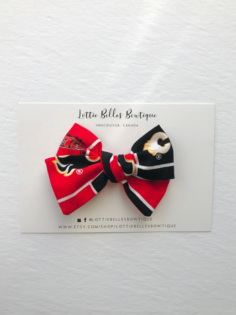 May include: A red, black, and yellow bow with a Calgary Flames and a  Edmonton Oilers hockey team logo. The bow is on a white card with the text "Lottie Belles Boutique, Vancouver, Canada" and the text "@lottie bellesboutique, www.etsy.com/shop/lottie bellesboutique".