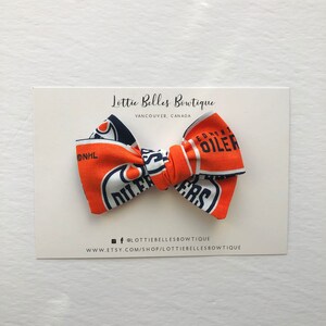 May include: A blue and orange bow tie with the Edmonton Oilers logo and NHL logo. The bow tie is on a white card with the text "Lottie Belles Boutique, Vancouver, Canada" and social media information.