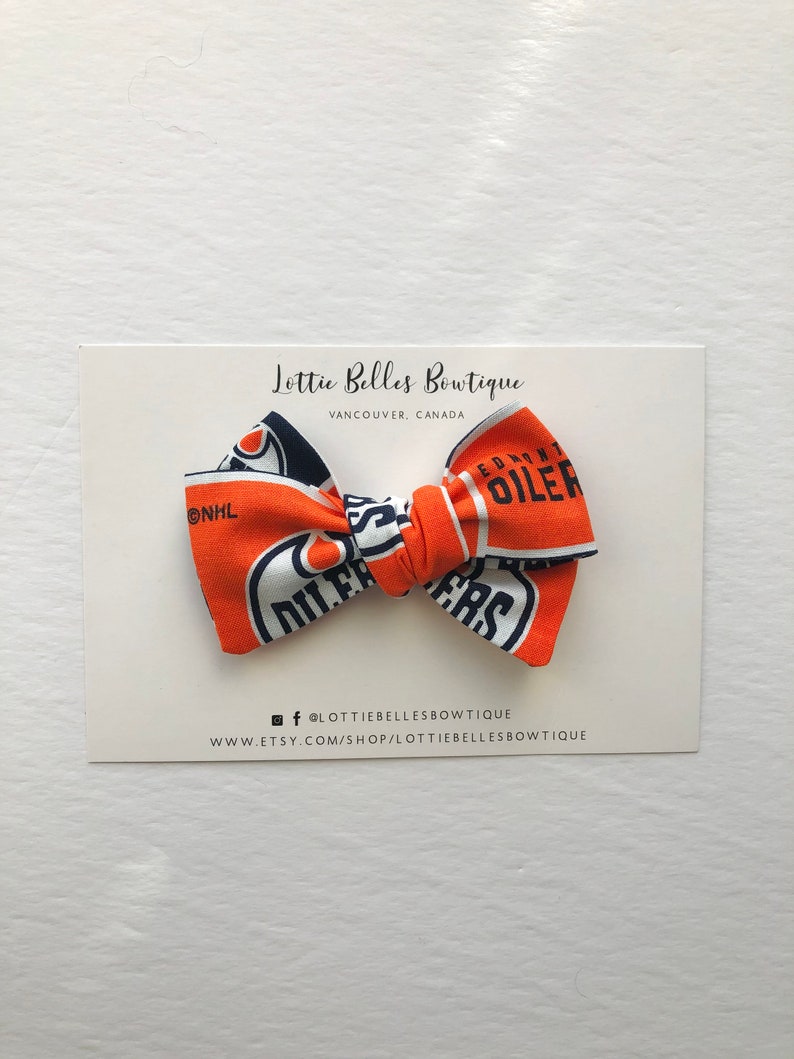 May include: A blue and orange bow tie with the Edmonton Oilers logo and NHL logo. The bow tie is on a white card with the text "Lottie Belles Boutique, Vancouver, Canada" and social media information.