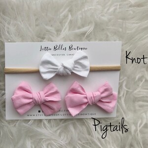 May include: A set of three hair bows, two pink and one white, with a nude elastic headband. The bows are tied in a knot and the headband is labelled "Lottie Belles Boutique" and "Knot Pigtails".