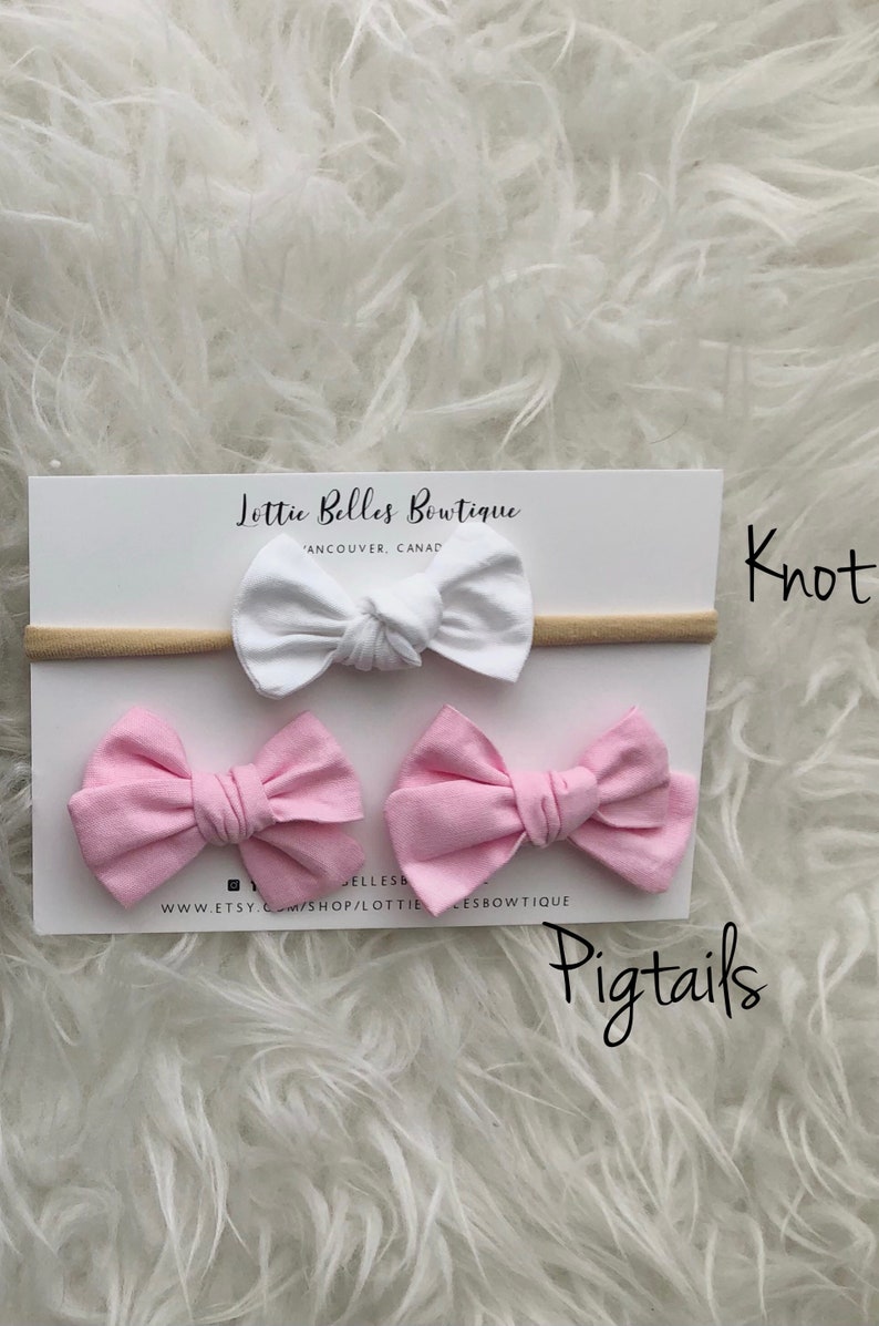 May include: A set of three hair bows, two pink and one white, with a nude elastic headband. The bows are tied in a knot and the headband is labelled "Lottie Belles Boutique" and "Knot Pigtails".