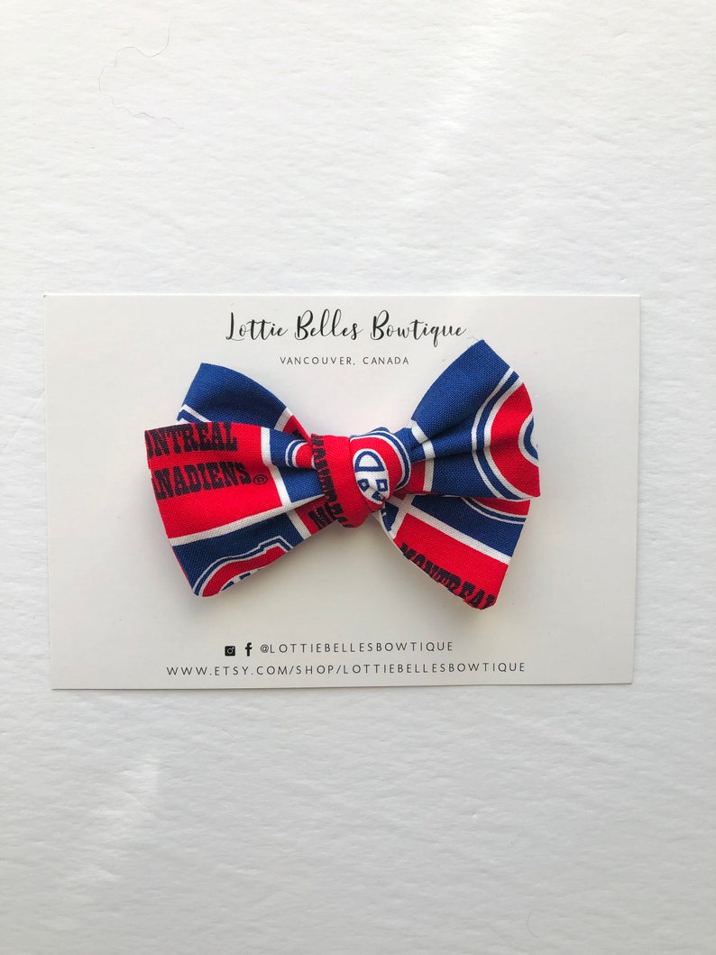 May include: A red and blue bow tie with the Montreal Canadiens logo. The bow tie is on a white card with the text "Lottie Belles Boutique, Vancouver, Canada" and social media information.