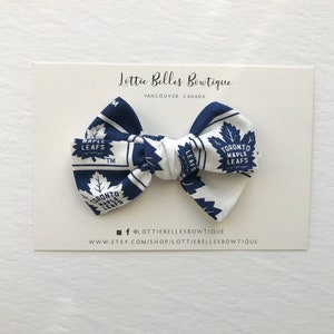 May include: A white and blue bow with a Toronto Maple Leafs pattern. The bow is on a white card with the text "Lottie Belles Bowtique" and "Vancouver, Canada".