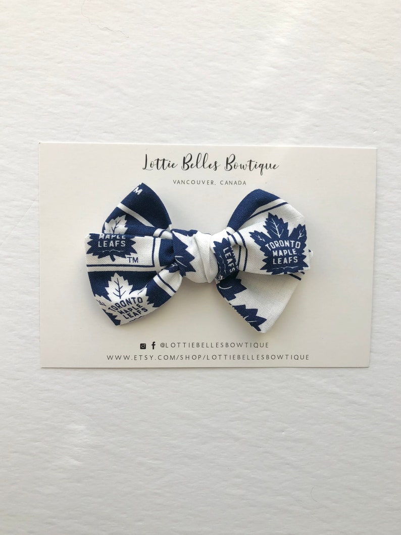 May include: A white and blue bow with a Toronto Maple Leafs pattern. The bow is on a white card with the text "Lottie Belles Bowtique" and "Vancouver, Canada".