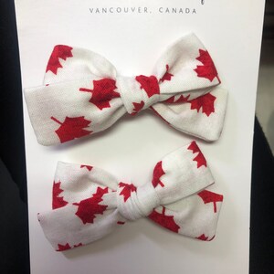 May include: Two white fabric hair bows with red maple leaf print. The bows are tied together with a white ribbon. The card reads "Lottie Belles Bowtique, Vancouver, Canada" and includes social media handles.