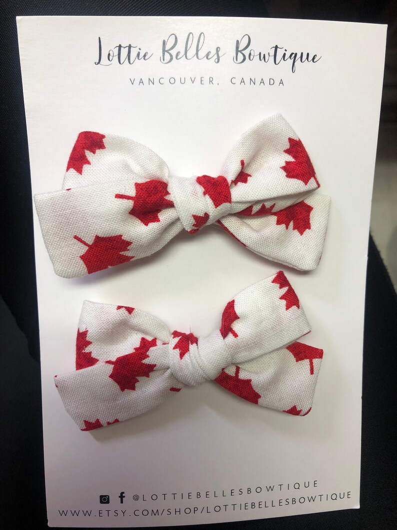 May include: Two white fabric hair bows with red maple leaf print. The bows are tied together with a white ribbon. The card reads "Lottie Belles Bowtique, Vancouver, Canada" and includes social media handles.