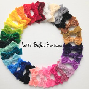 May include: A vibrant array of velvet hair bows in a circular arrangement, showcasing a spectrum of colours. The bows include shades of red, orange, yellow, green, blue, and purple. The text "Lottie Belles Boutiquie" is visible.