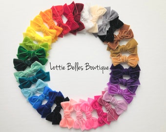 May include: A vibrant array of velvet hair bows in a circular arrangement, showcasing a spectrum of colours. The bows include shades of red, orange, yellow, green, blue, and purple. The text "Lottie Belles Boutiquie" is visible.
