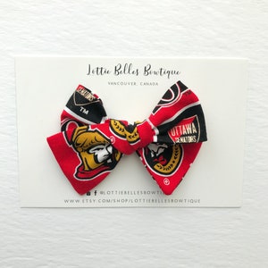 May include: A red, black, and gold bow with the Ottawa Senators logo. The bow is made of fabric and has a white background. The bow is tied with a white ribbon. The bow is on a white card with the text "Lottie Belles Boutique" and "Vancouver, Canada".