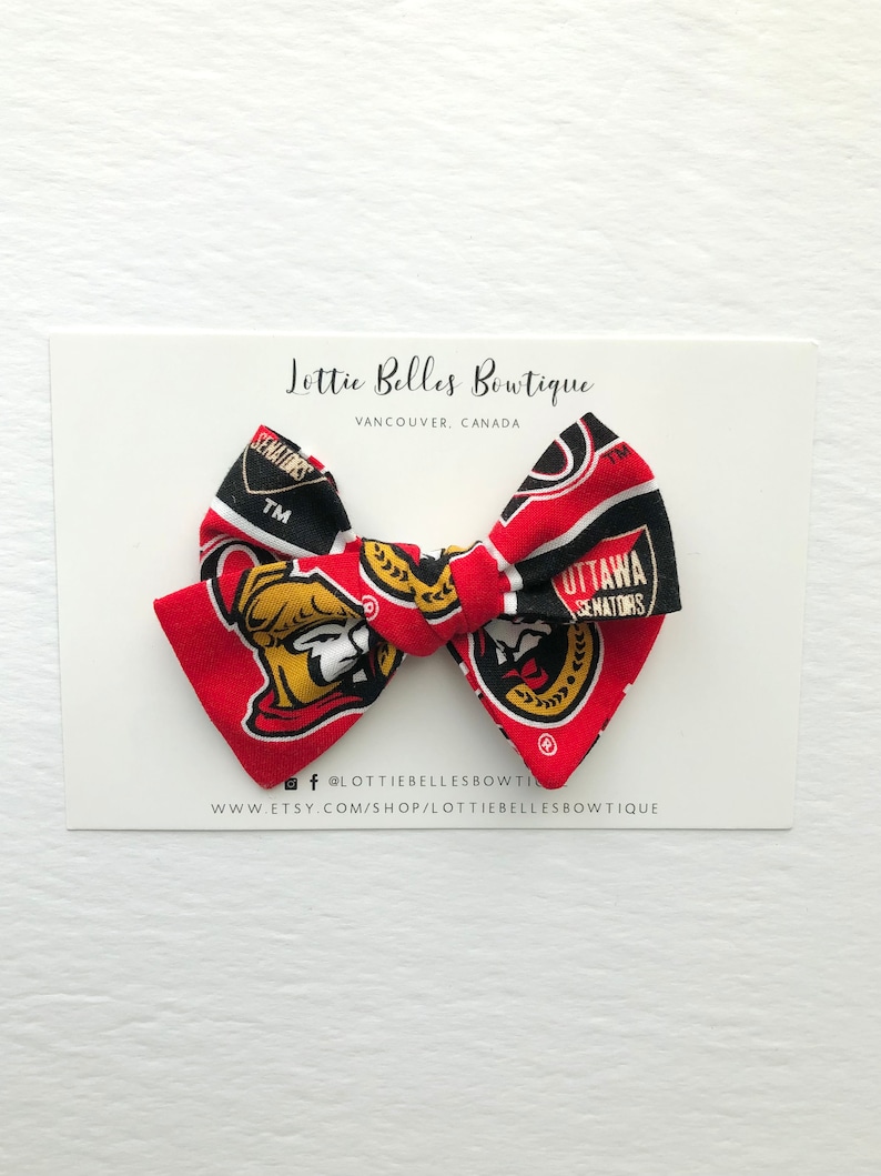 May include: A red, black, and gold bow with the Ottawa Senators logo. The bow is made of fabric and has a white background. The bow is tied with a white ribbon. The bow is on a white card with the text "Lottie Belles Boutique" and "Vancouver, Canada".