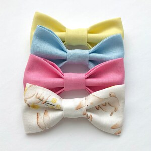 May include: Three fabric bow ties. The top bow tie is yellow, the middle bow tie is light blue, and the bottom bow tie is pink. The bottom bow tie has a white background with a pattern of brown bunnies and yellow flowers.