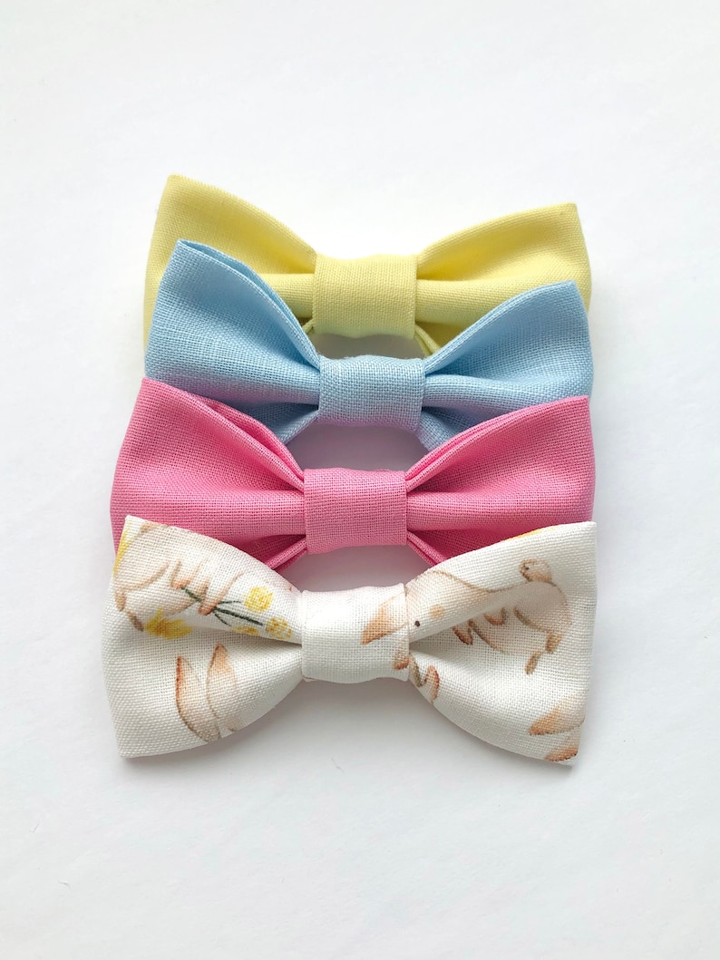 May include: Three fabric bow ties. The top bow tie is yellow, the middle bow tie is light blue, and the bottom bow tie is pink. The bottom bow tie has a white background with a pattern of brown bunnies and yellow flowers.